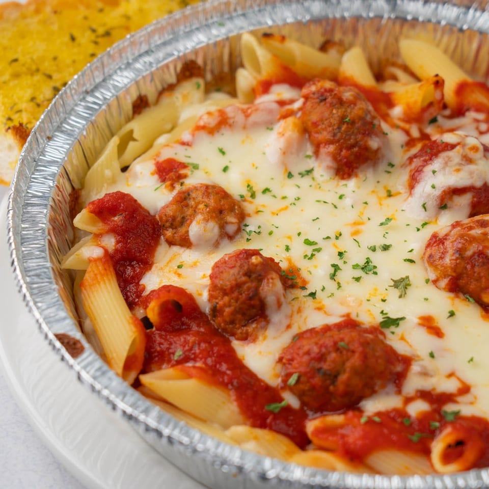 Baked penne pasta with meatballs, melted mozzarella, marinara sauce, and parsley in a foil container.