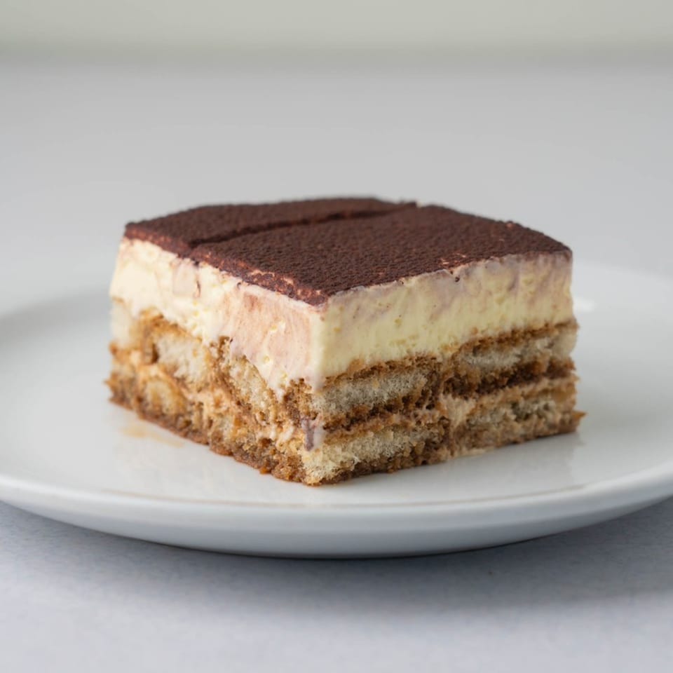 A slice of tiramisu with layers of ladyfingers, cream, and cocoa powder on a white plate.