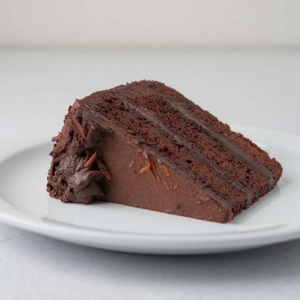 A rich slice of TJ's chocolate cake with chocolate frosting and shavings on a white plate.